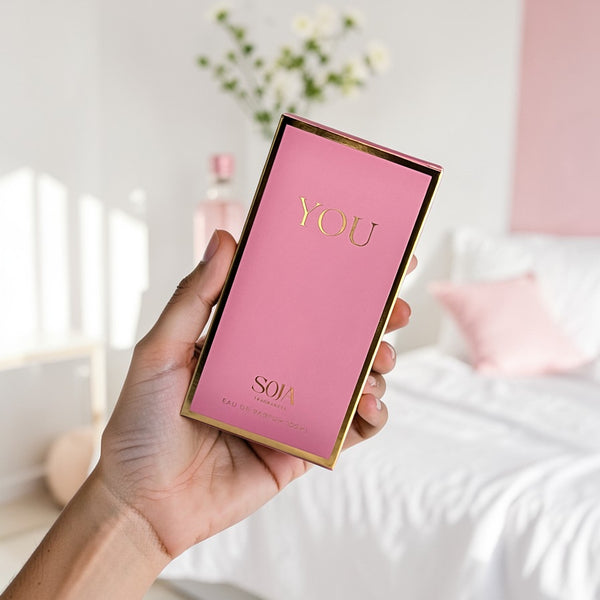 Hand holding a pink and gold product box with 'YOU' and 'Soja' text in a blurred indoor setting.