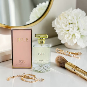 Perfume bottle and packaging labeled 'True' on a surface with flowers and jewelry.
