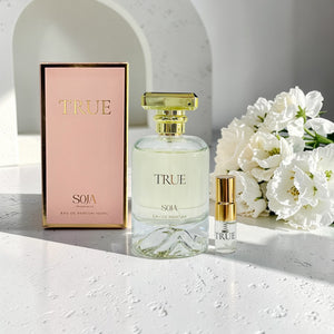 True Soja perfume bottle and packaging on a light surface with flowers in the background