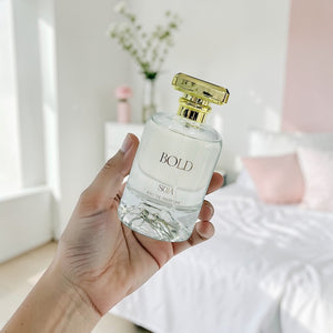 Hand holding a perfume bottle labeled 'BOLD' with a blurred indoor background