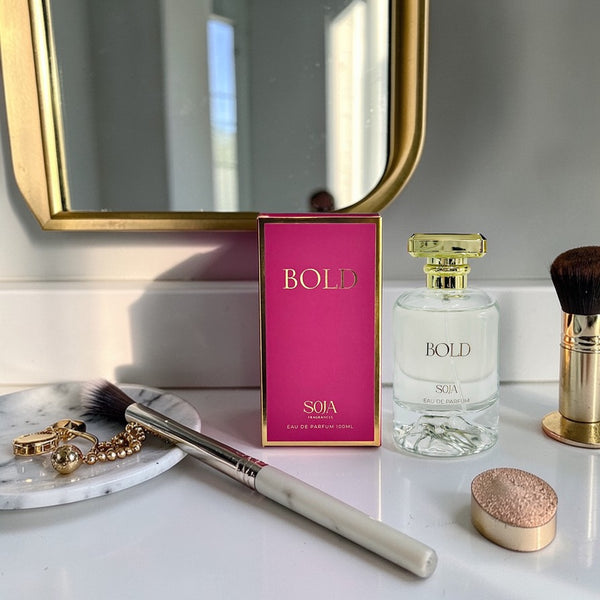 Perfume bottle, packaging, makeup brush, and jewelry on a vanity table with a mirror in the background.
