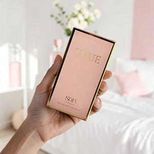 Hand holding a pink and gold 'True' perfume box with a blurred indoor background