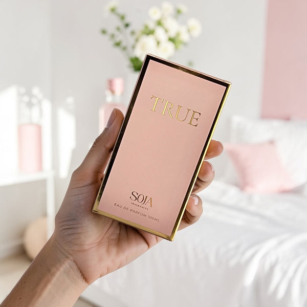 Hand holding a pink and gold 'True' perfume box with a blurred indoor background