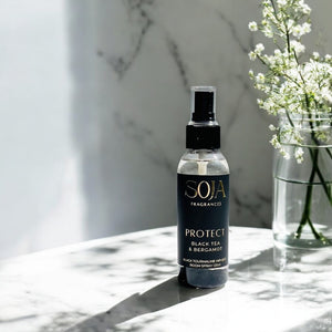 Spray bottle labeled 'SOJA Fragrances' on a marble surface with a vase of flowers in the background.