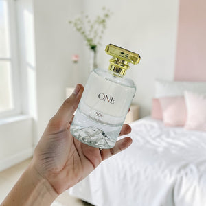 Hand holding a clear perfume bottle with gold cap in a softly lit room.