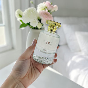 Hand holding a perfume bottle labeled 'YOU' with a blurred background of flowers and a bed.