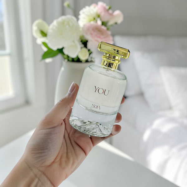 Hand holding a perfume bottle labeled 'YOU' with a blurred background of flowers and a bed.