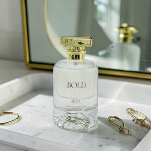 Clear perfume bottle with gold cap labeled 'BOLD' on a marble surface with jewelry.