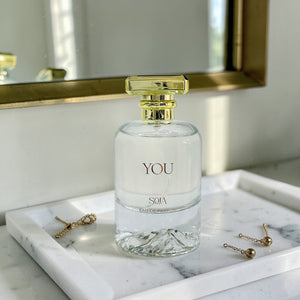 Perfume bottle labeled 'YOU' by 'SOJA' on a marble surface with jewelry.